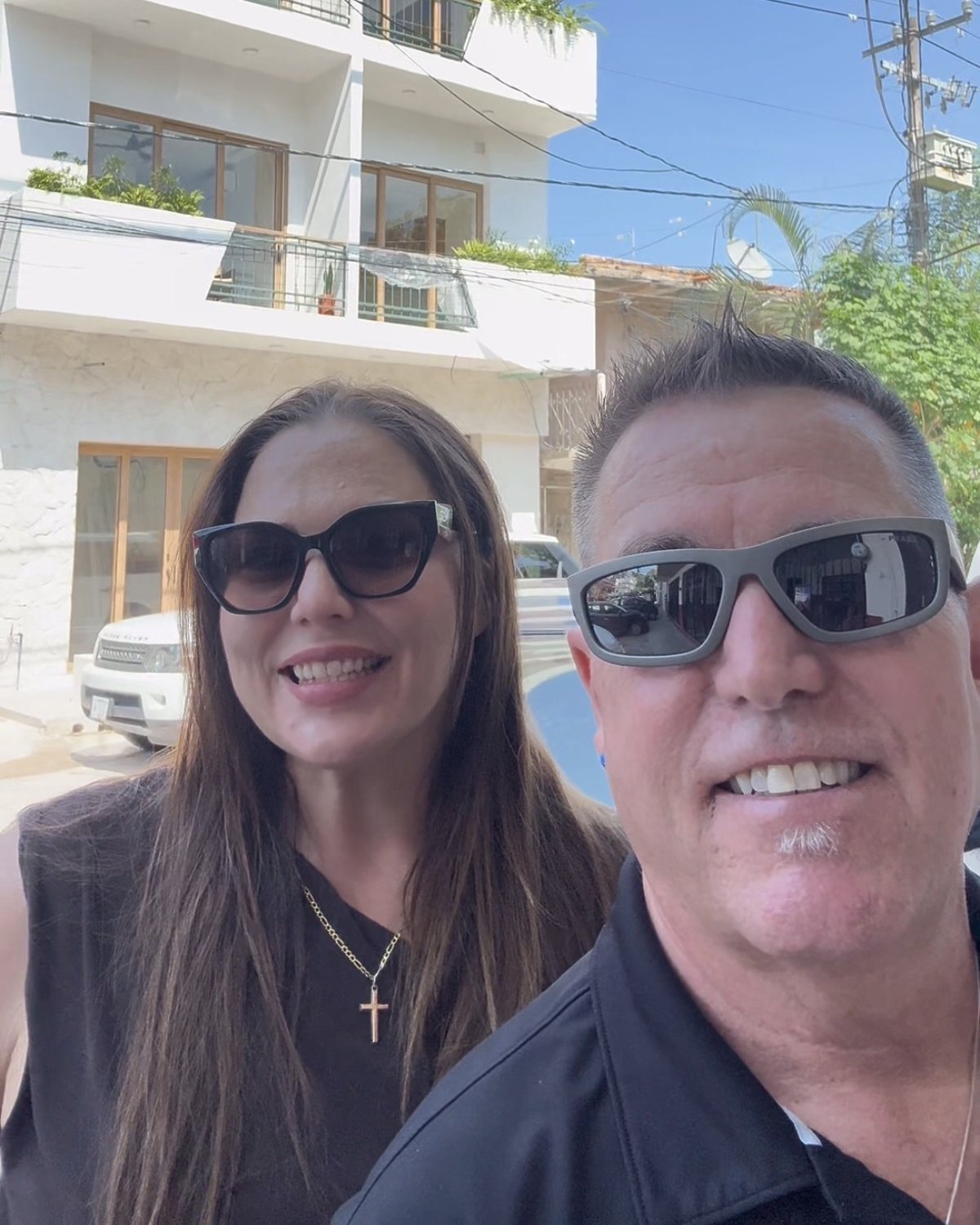 Hannah L&oacute;pez and Tom Gordon smiling in sunglasses in front of Tr&oacute;pico 1371, a white condo building in Puerto Vallarta with wood-framed windows, balconies and planters