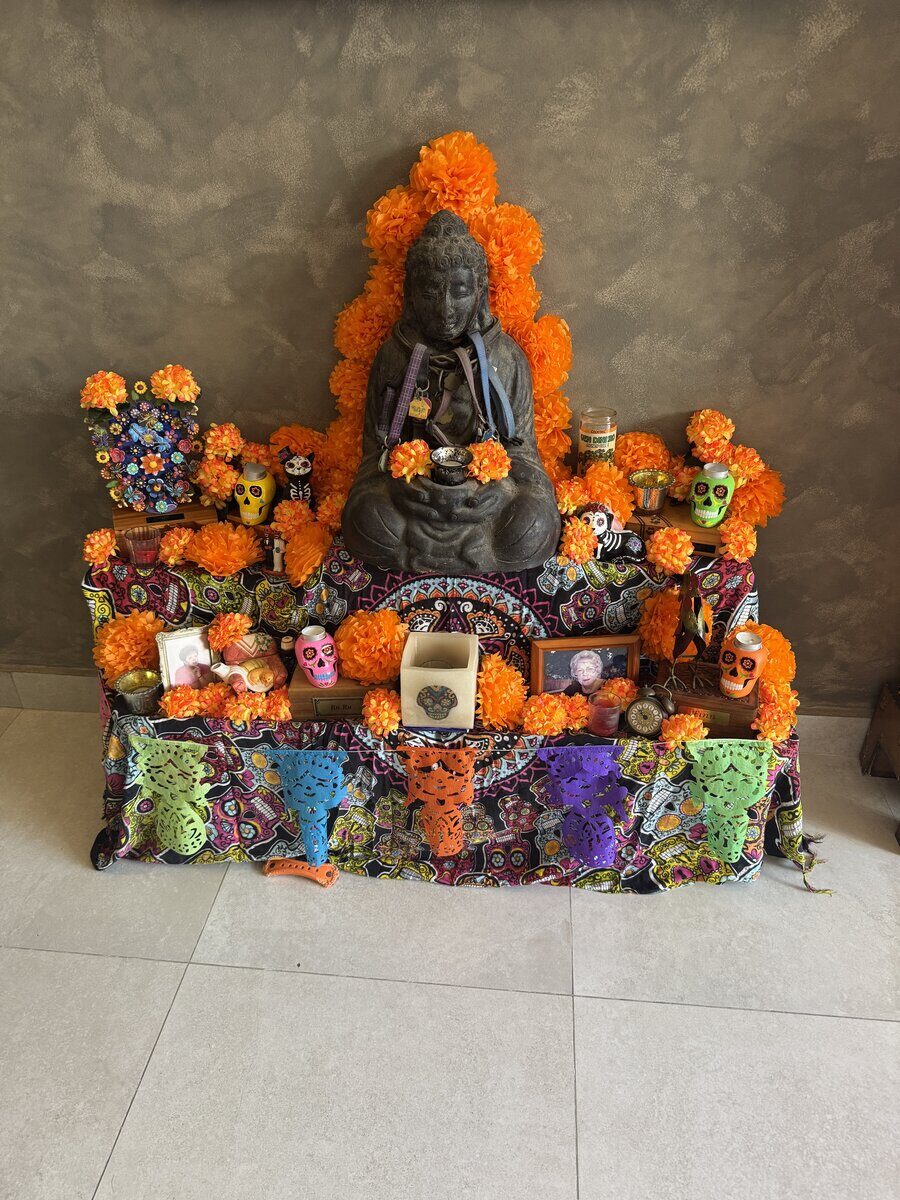 A small Buddha figure at the base of an ofrenda with marigolds, sugar skulls and a lit candle