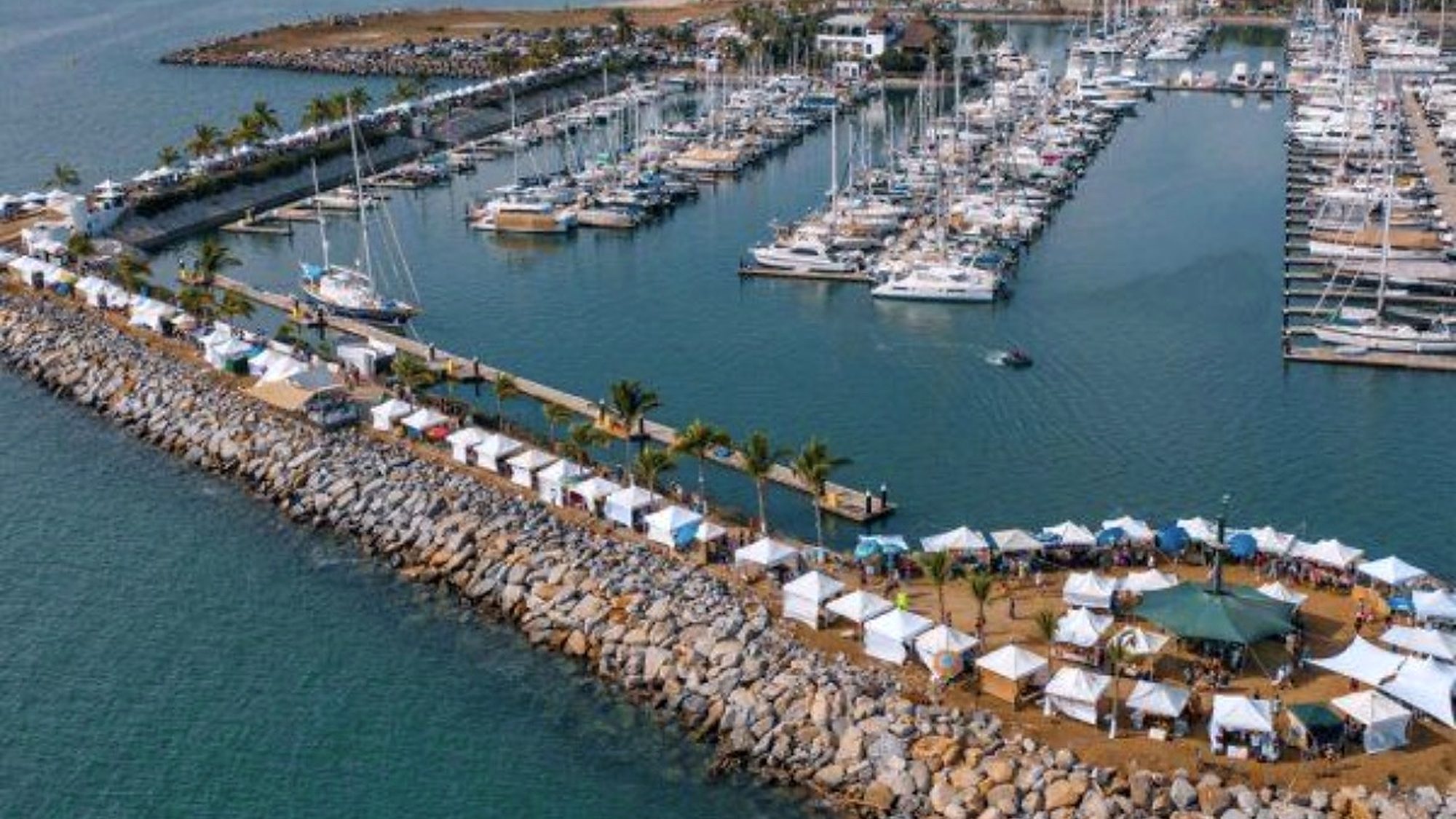 Aerial view of the Sunday market stalls along the breakwater at the La Cruz marina, with boats in the slips and the hills of the north shore behind