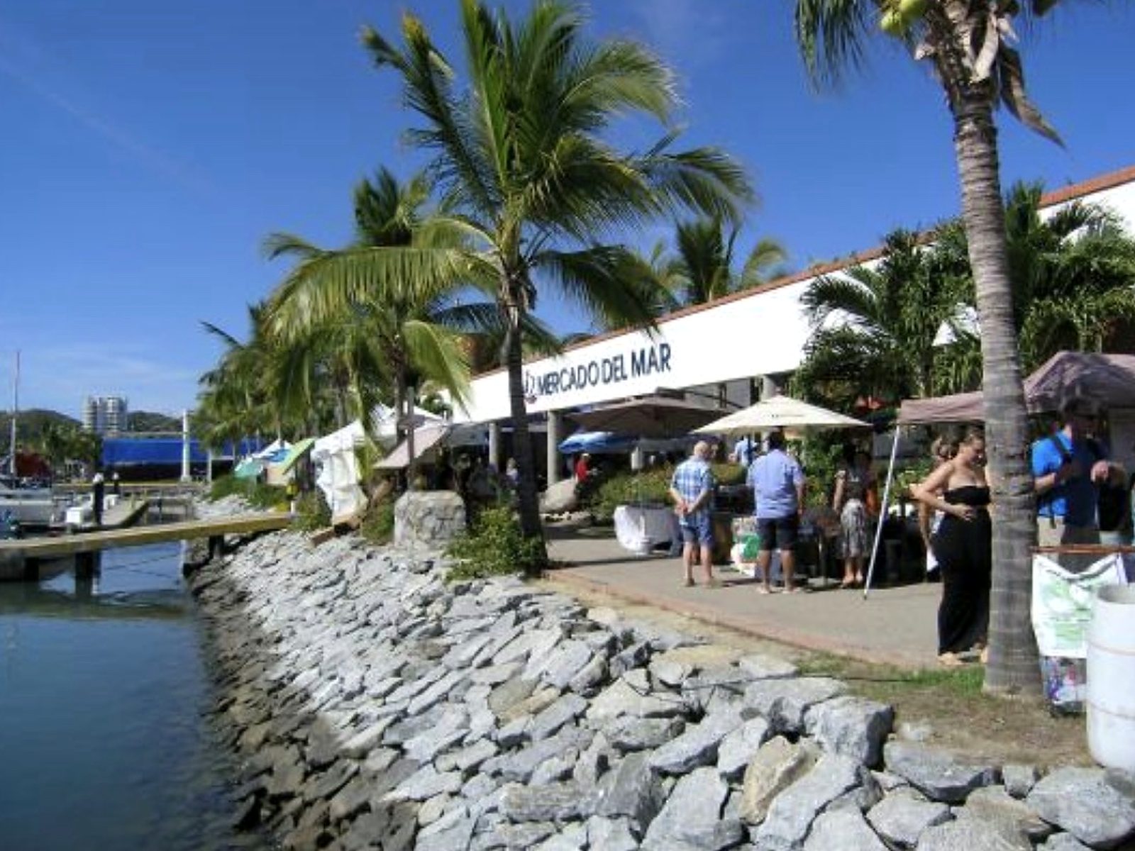 The Mercado del Mar fish market building at the La Cruz marina, with palms, shoppers and a rock seawall