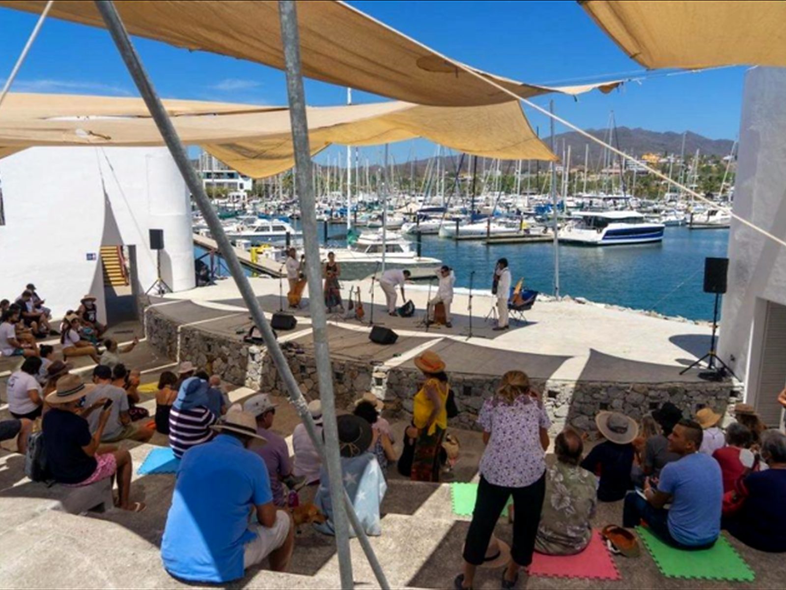 A crowd seated under shade sails watching live music at the La Cruz marina, with sailboats in the slips behind
