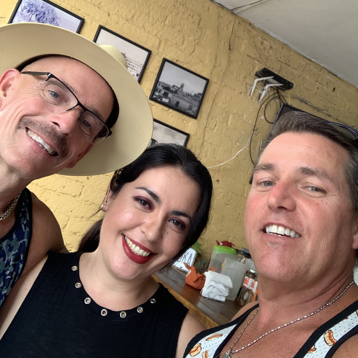 Chuck, Karla and Tom smiling in a selfie against a yellow brick wall hung with black and white photographs